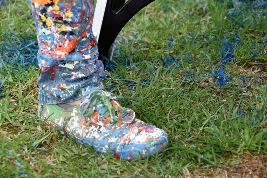 An artists' boots and trousers cuff covered in splashes of paint as they stand on lawn.