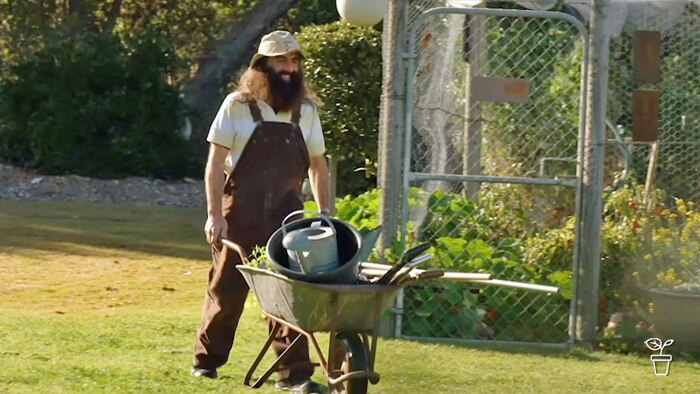 Man with hat and a beard pushing a wheelbarrow filled with gardening tools through a garden