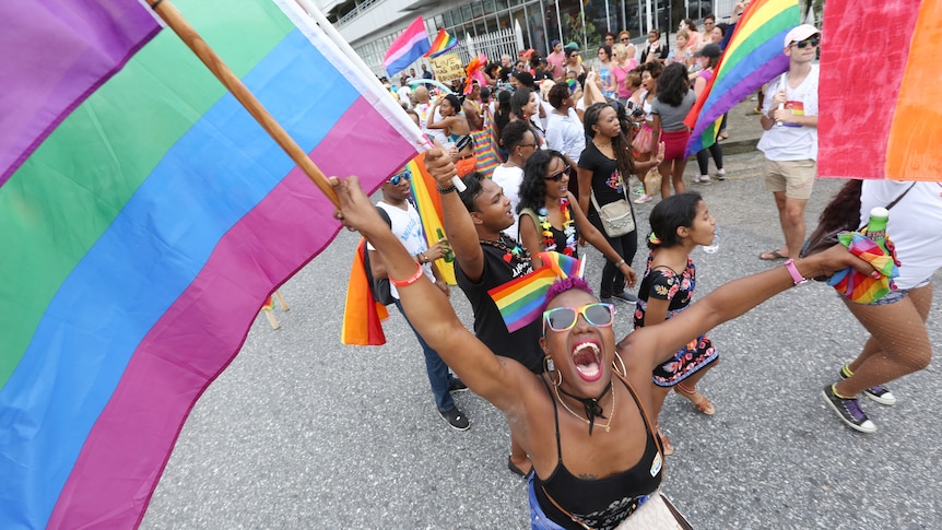 A group of people walk down a street holding rainbow flags. The closest to the camera makes eye contact, smiling widely