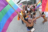 A group of people walk down a street holding rainbow flags. The closest to the camera makes eye contact, smiling widely