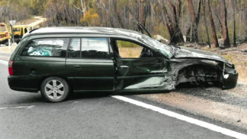 A smashed car lies across a road, with the front door and wheel ripped off