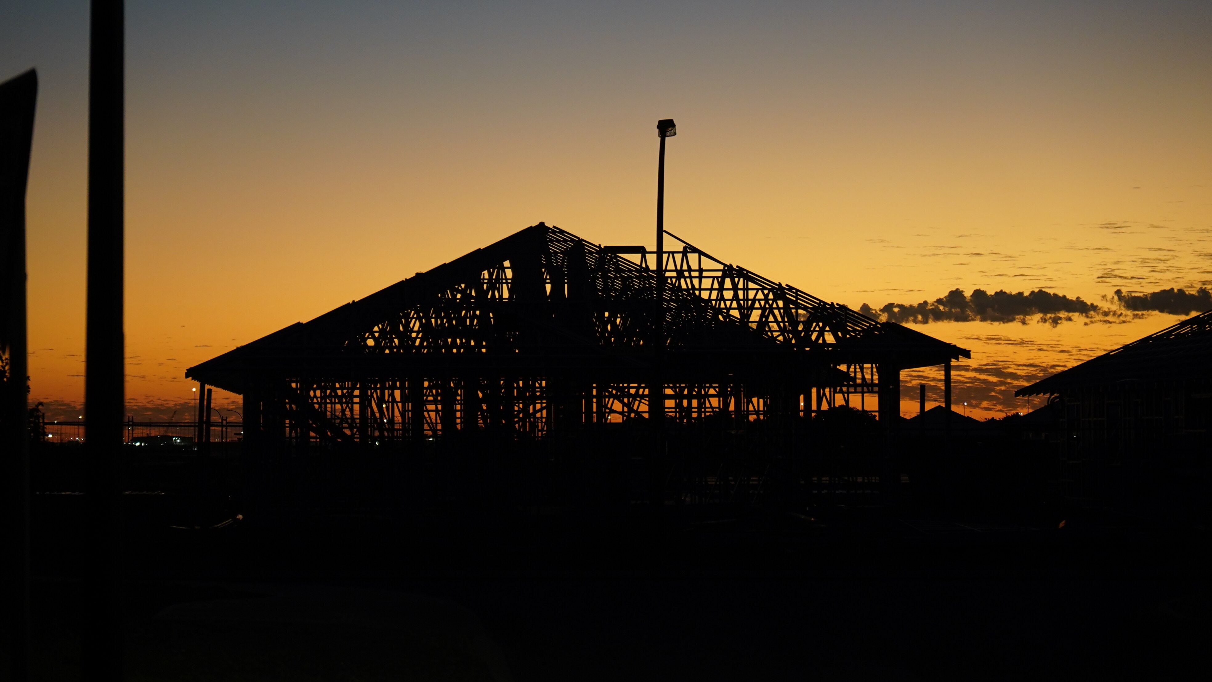 The steel frame of a house in mid-construction, in front of a light orange sunset.