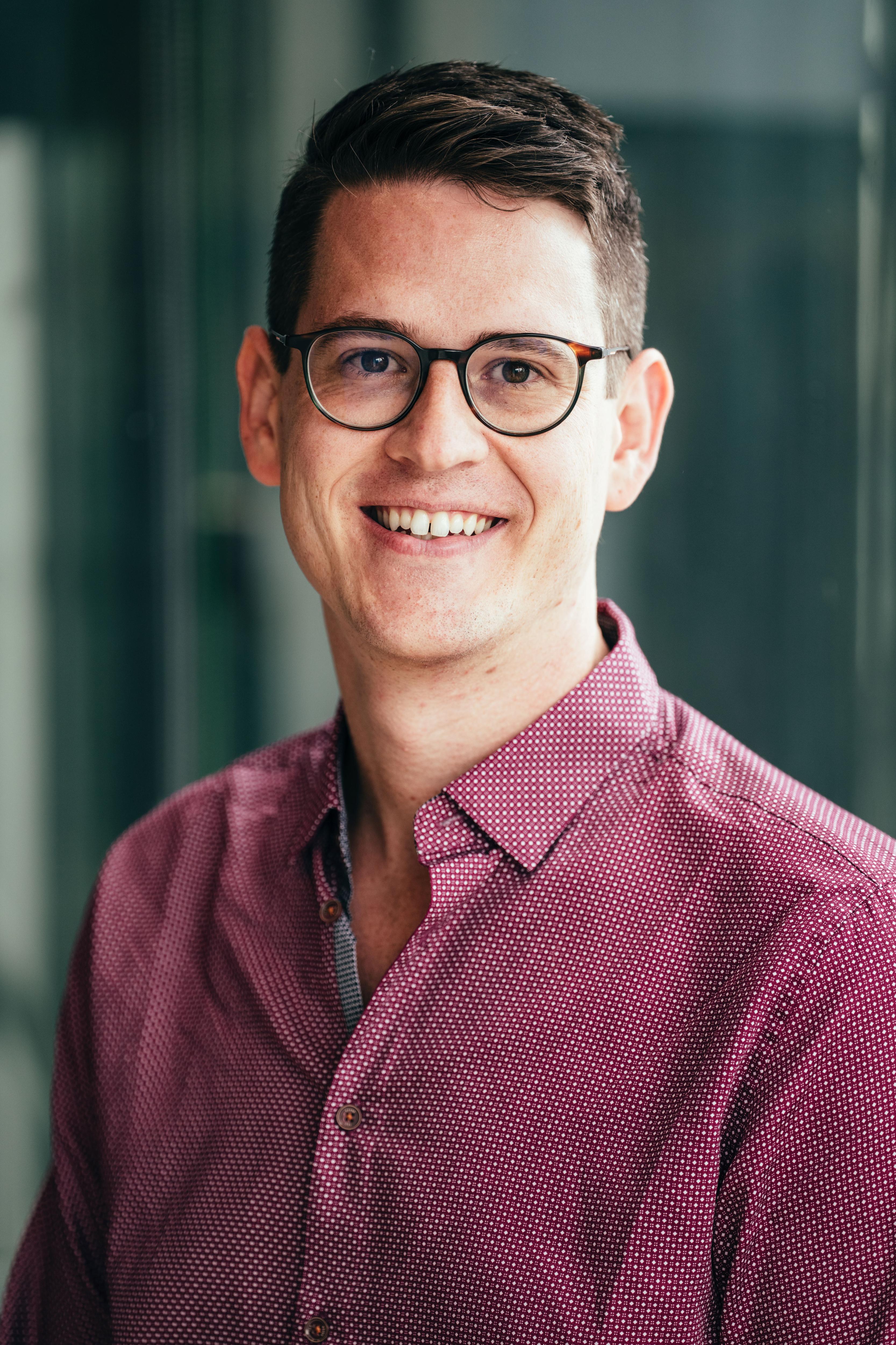 A man wearing a dark red shirt and black-framed glasses smiles at the camera.