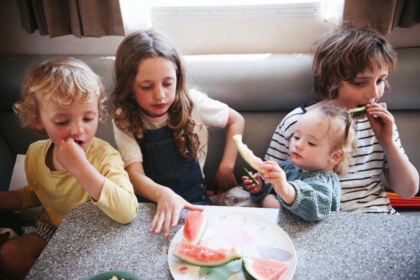 Four young children sit on a bench at a small table eating watermelon.