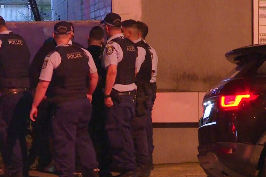 A number of police with backs to the camera next to a car at night.