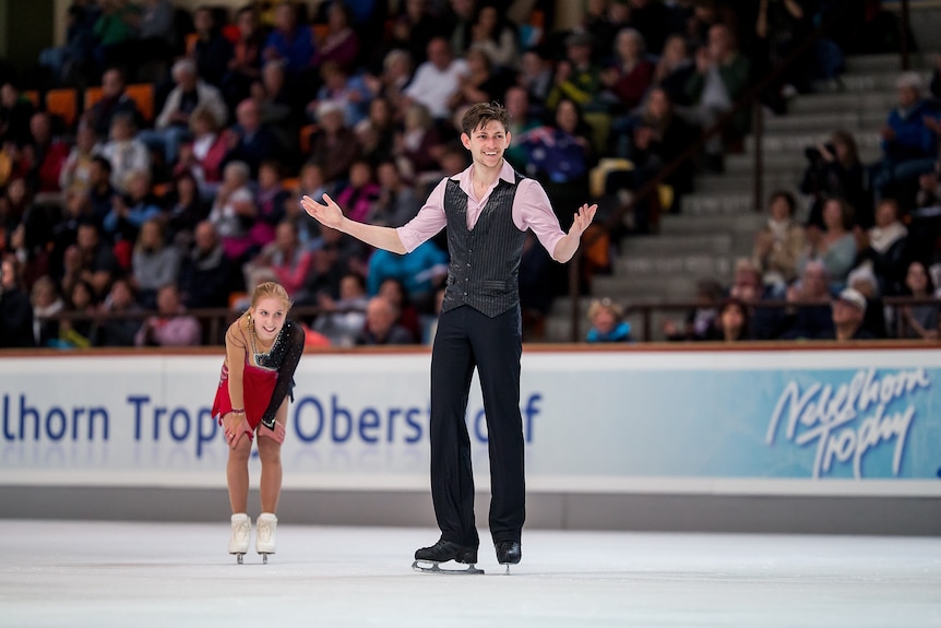 A figure skating pair react - the male skater smiles and holds his hands out in surprise, while the female skater grins.