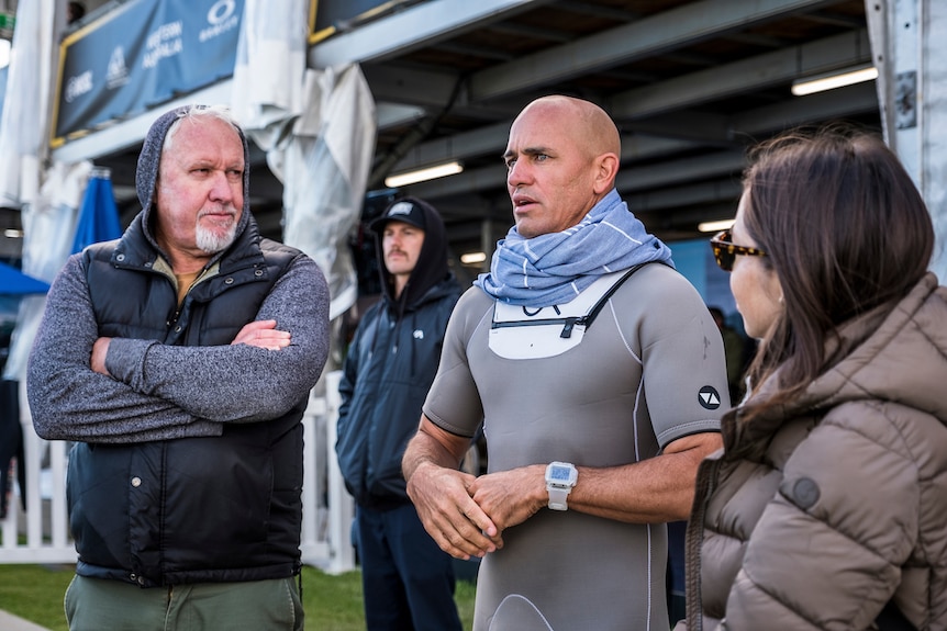 Thorson stands next to Slater in a wetsuit who is beside a woman in a puffer jacket, talking seriously as Slater looks ahead.