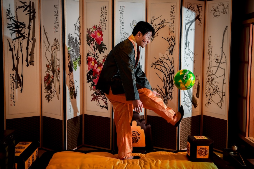 A man wearing traditional Korean clothes juggles a green and gold football in a traditional Korean house.
