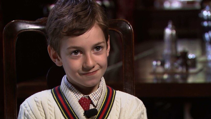 A young boy in a shirt, tie and jumper sits in a chair.