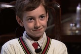 A young boy in a shirt, tie and jumper sits in a chair.