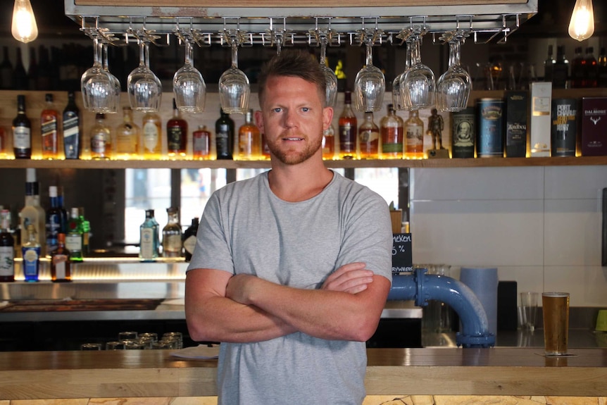Sandbar owner Ben Randall stands with his arms folded in front of a bar.