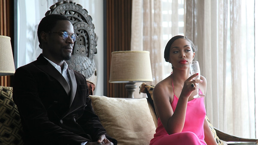 Indoors, man with round glasses wears velvet black suit and sits next to woman in long formal magenta dress holding tall glass.