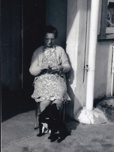 A black and white photo shows a woman sitting in a chair knitting, circa 1976.