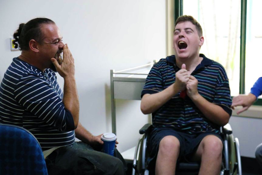 A man holds his hand over his face laughing as a man in a wheelchair has a big laugh
