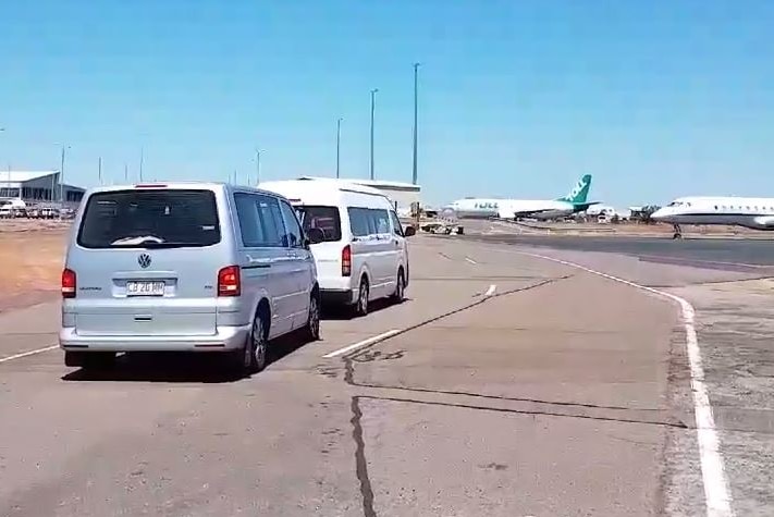 Two white vans driving on a road with two planes in the background.
