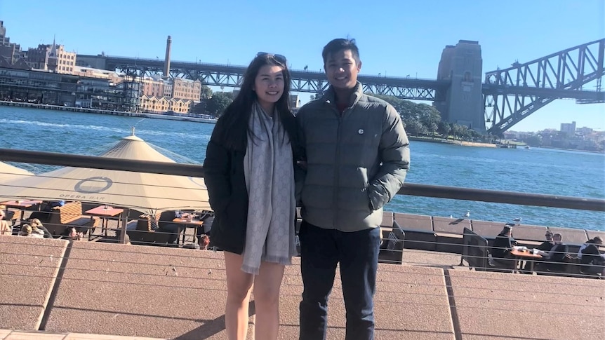Couple stand looking at camera with Sydney Harbour behind them. 