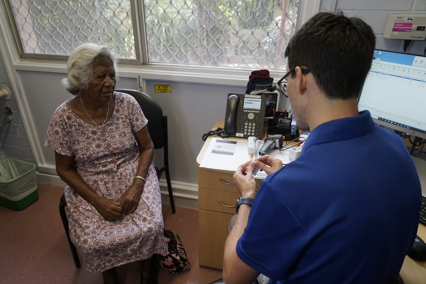 An elderly Indigenous lady sits in a doctors room waiting to receive a vaccination.  