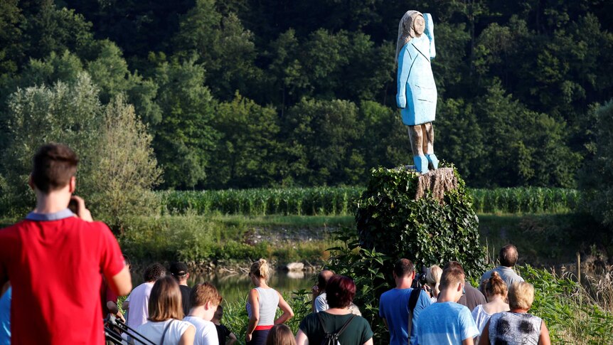 A crowd of people stand at the base of a tree, which has been carved into the shape of a woman, in a painted blue dress.