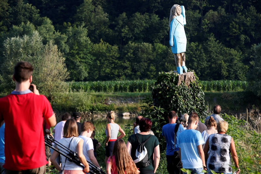 A crowd of people stand at the base of a tree, which has been carved into the shape of a woman, in a painted blue dress.