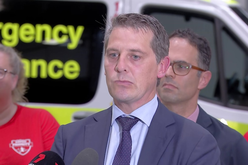 Ryan Park in suit stands in front of an ambulance behind a row of microphones