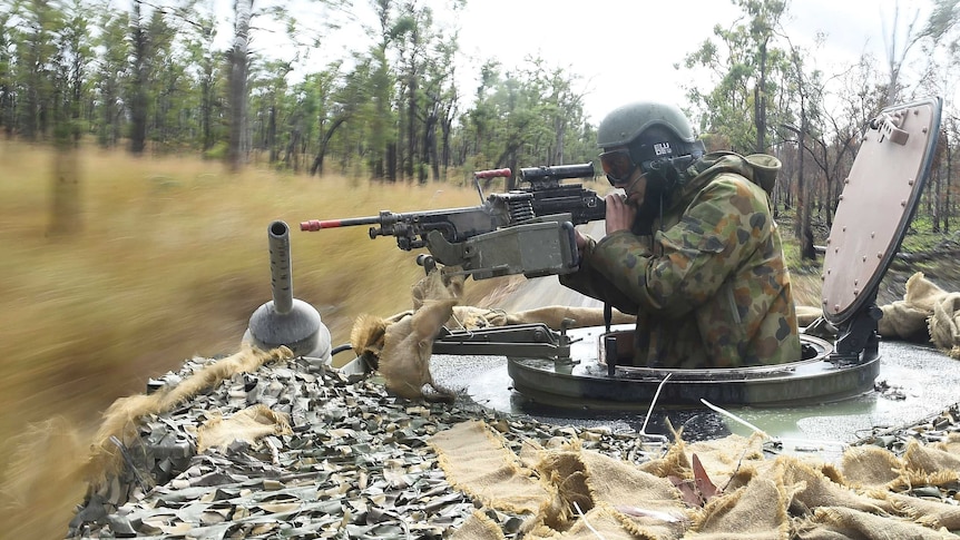 A solider with a machine gun, sitting in a moving tank, looks into the bushes.