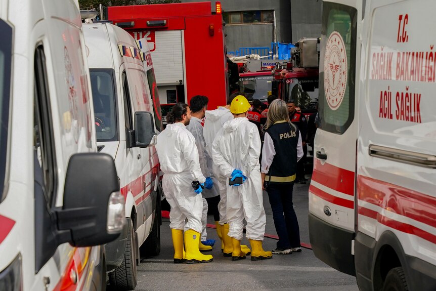 Emergency service workers in white overalls and yellow boots stand between ambulances on a street