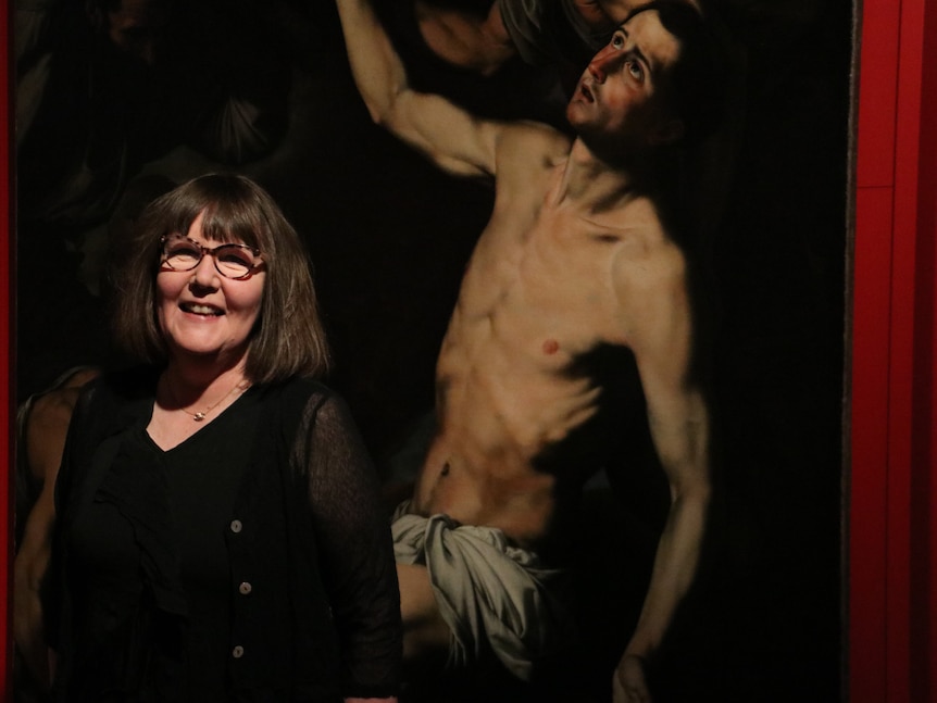 A woman stands in front of historic painting with male human figure in loin cloth