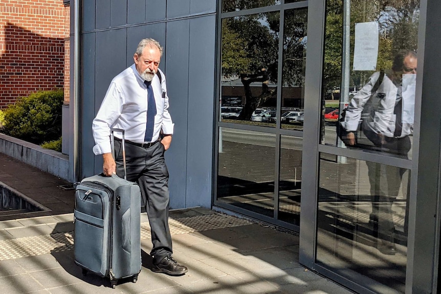 An elderly man looks at the camera as he enters court carrying a suitcase.