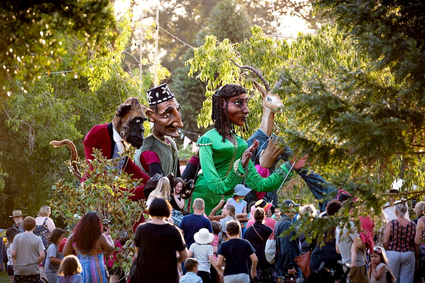 Three large puppets move through some trees and into a crowd.