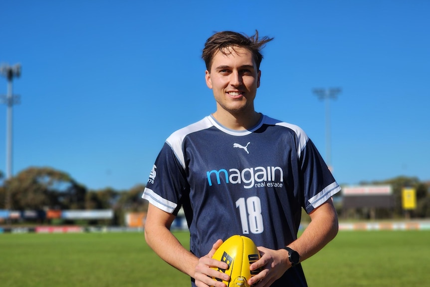 A man in a blue jersey holding a yellow football smiling on a sunny day.