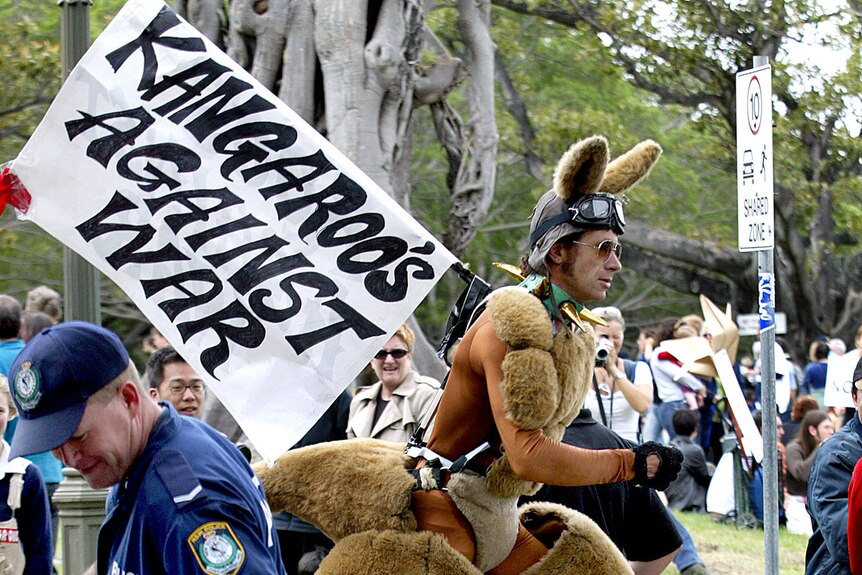 A man dresses as a kangaroo carries a sign that says "Kangaroos against war" as he marches through a rally in a park