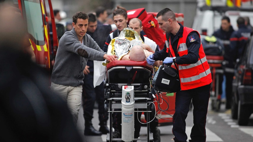 A patient is carried on a stretcher by paramedics as other emergency services personnel work in the background.