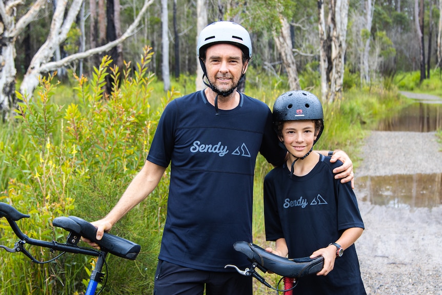 A father stands with his arm around his son, as they both hold unicycles and wear helmets on a bush track.