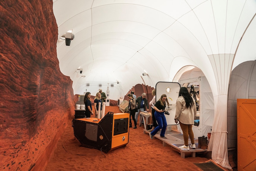 A space with red sand and printed red rocks is pictured. People with cameras are walking through it.