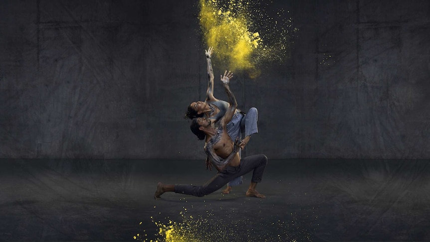 A male and female dancer in a grey studio throw ochre-coloured dust into the air.