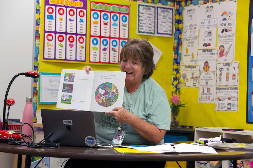 A woman holds up a storybook to a computer in a classroom