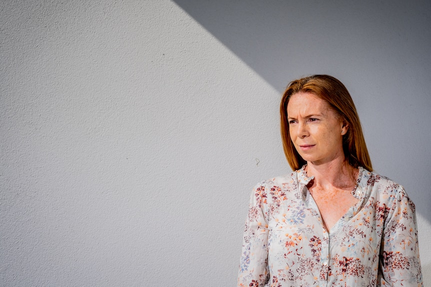 a woman wearing a top and jeans stands against a white wall that's highlighted horizontally by natural light