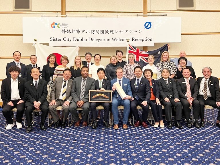A group of men and women sit posing for a photo below a sign that says Sister City Dubbo Delegation Welcome Reception.