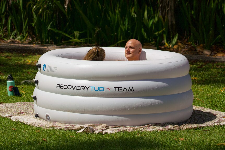  Two people sit in a white ice bath, which is on green grass. They lay back and relax.