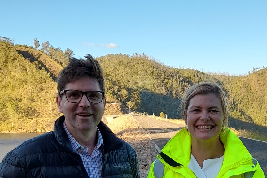 A dark-haired man in a puffer jacket stands next to a smiling blonde woman in high-vis. They are in front of a dam.