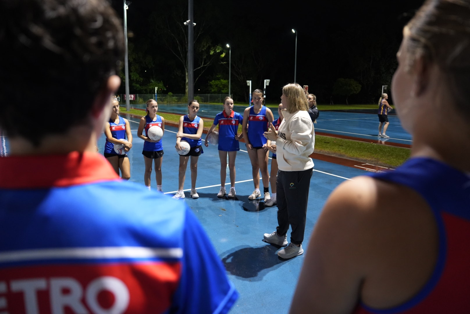Stacey Marinkovich talks to young netball players who are standing around in a huddle listening on the netball courts.