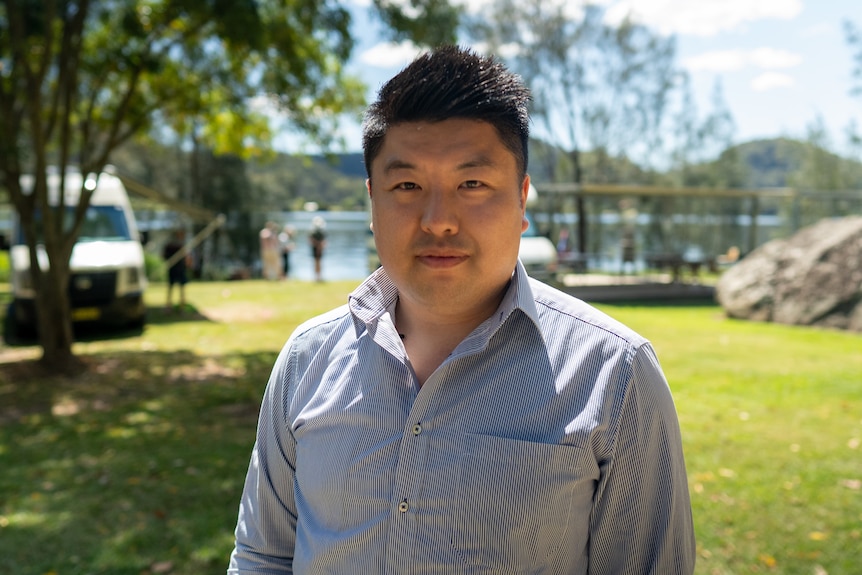 A man stands outside with grass and water in the background.