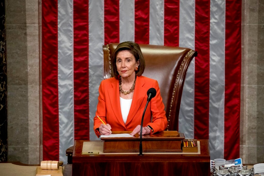 Democratic House Speaker Nancy Pelosi stands in front of the speaker's chair in front of a large American flag.