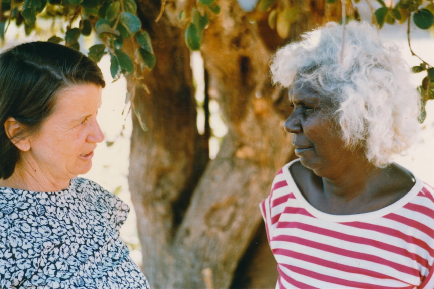 A caucasian woman and indigenous stand side by side and are smiling at each other.