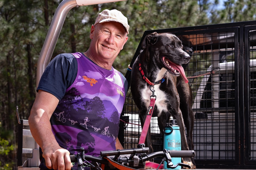 A smiling, middle-aged man stands near his dog, which is on the back of a ute.
