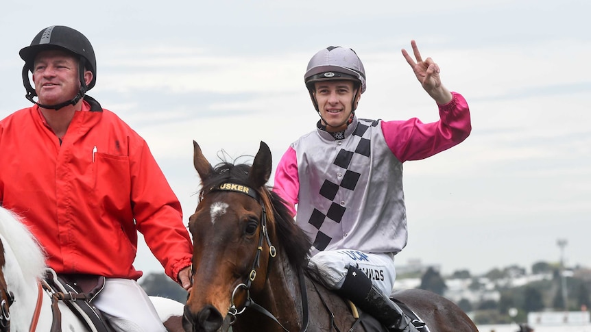 A jockey throws up the peace sign while riding a horse.