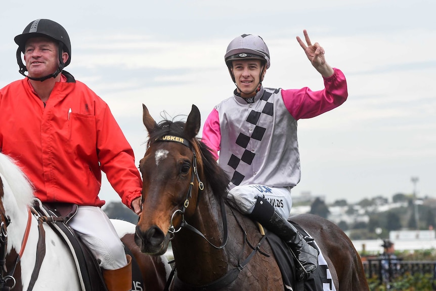 A jockey throws up the peace sign while riding a horse.