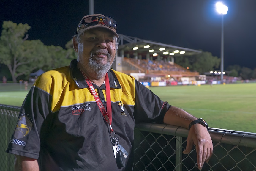 A man smiles at the camera while leaning on a fence next to a footy field