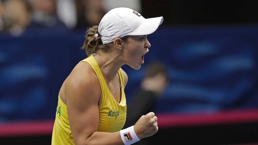 Ash Barty clenches her fist and yells while wearing a white baseball cap and yellow singlet