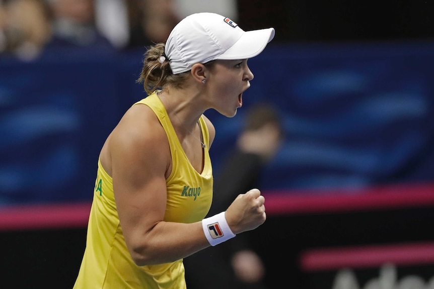 Ash Barty clenches her fist and yells while wearing a white baseball cap and yellow singlet
